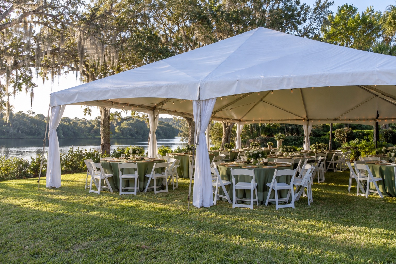 Family riverside-style banquet on lawn, white canopy with sidewalls rolled up, circular tables with green linens, white chairs, wooded backdrop, morning