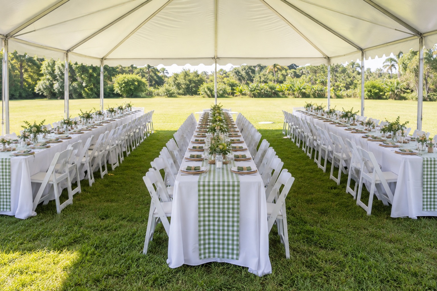 Large family gathering with three parallel banquet table rows under white marquee, gingham sage runners and white folding chairs, open grass field, bright afternoon