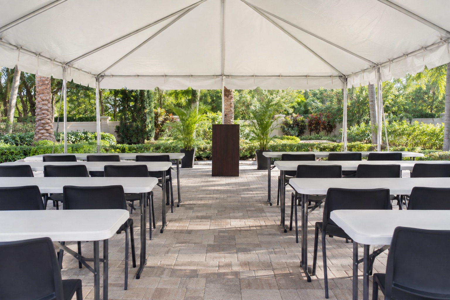 Professional outdoor meeting layout beneath white frame tent, classroom rows of narrow white tables and black chairs, simple unbranded podium, garden courtyard, morning