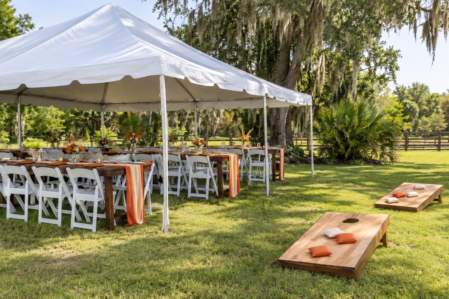Relaxed rural backyard birthday setup with white canopy, orange and cream table settings on wooden tables, white folding chairs, separate lawn cornhole boards, daytime