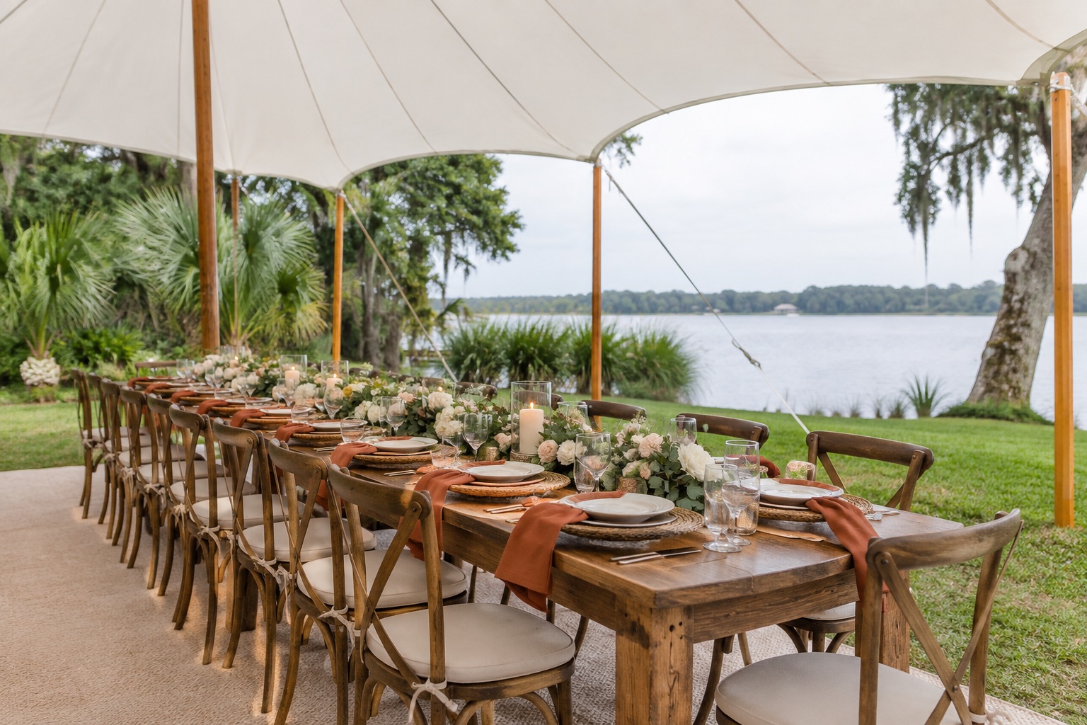 Intimate outdoor wedding reception, one long wooden banquet table under a small sailcloth tent, terracotta napkins and crossback chairs, soft lake view, overcast luminous daylight