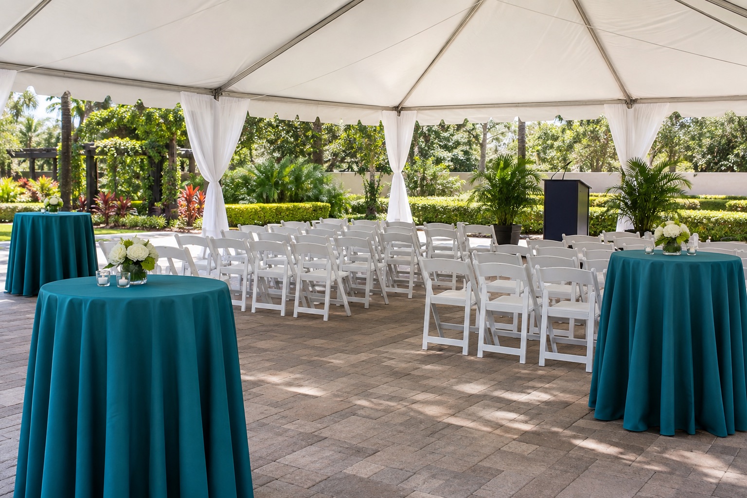School or company celebration in a garden courtyard under white tent, teal cocktail tables alongside rows of white chairs and a modest blank podium, bright afternoon